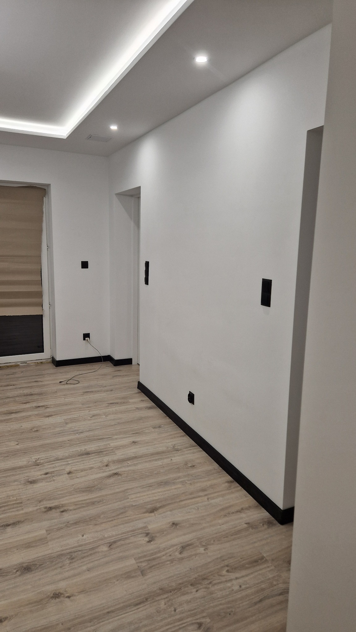 Wnętrze po remoncie z białymi ścianami, drewnopodobną podłogą i czarnymi listwami przypodłogowymi oraz gniazdkami; widoczne oświetlenie sufitowe LED.