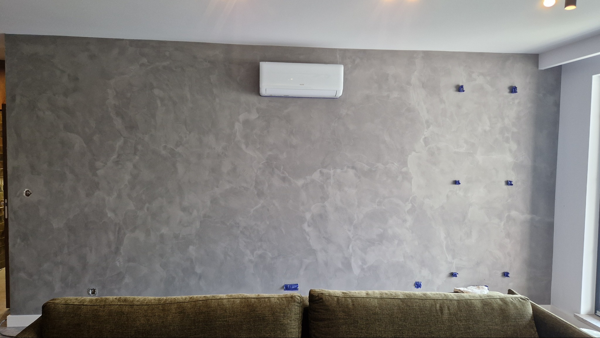 Ściana z dekoracyjnym tynkiem w odcieniach szarości, z widocznym klimatyzatorem i niebieskimi elementami montażowymi, częściowo zasłonięta przez górną część sofy.