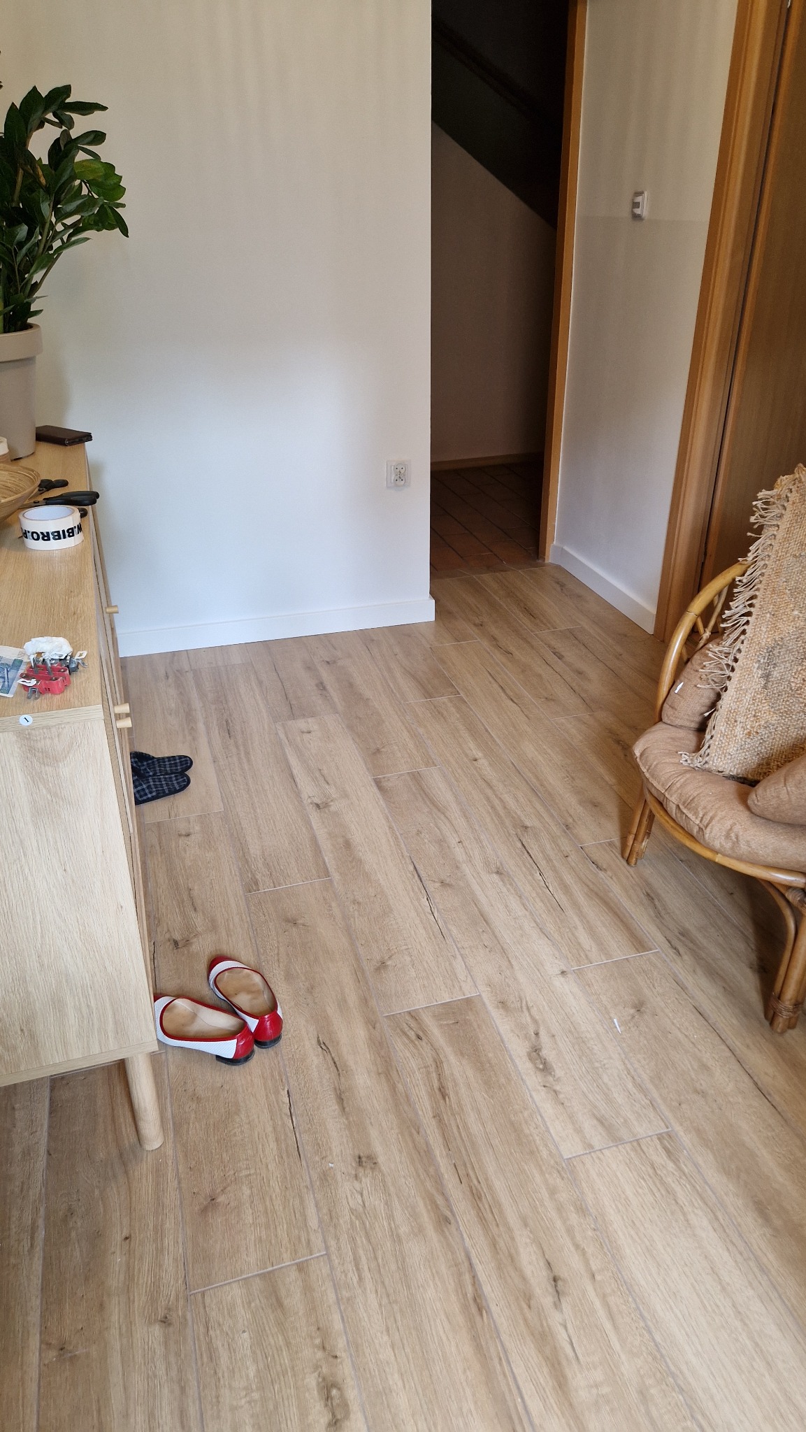 Jasne, drewnopodobne płytki podłogowe ułożone w korytarzu, widoczne buty damskie z czerwonymi elementami, rattanowy fotel z poduszką i fragment szafki z rośliną doniczkową.