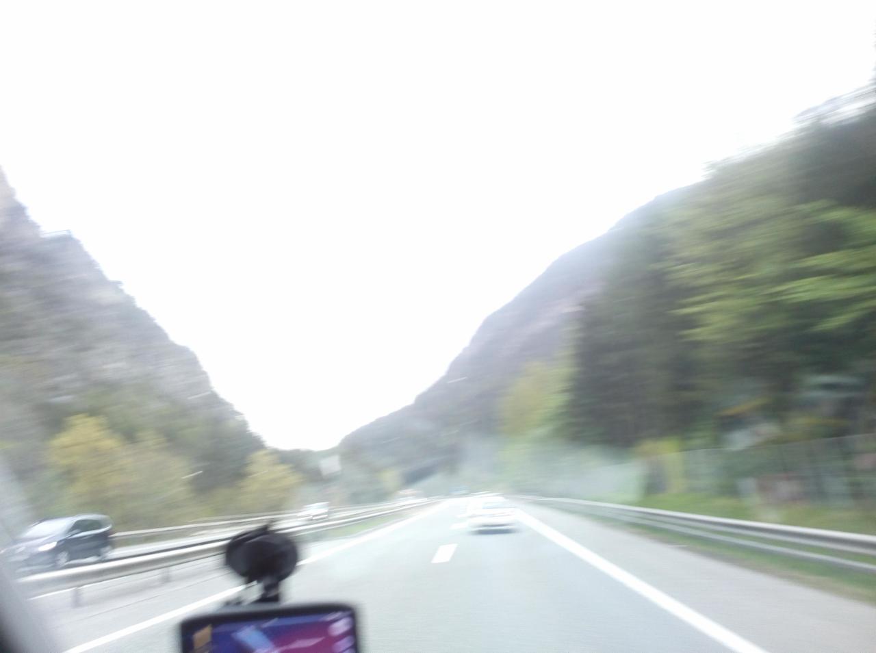 Widok z wnętrza pojazdu na autostradę prowadzącą przez górzysty teren; widoczny czarny uchwyt na telefon i fragment nawigacji GPS.