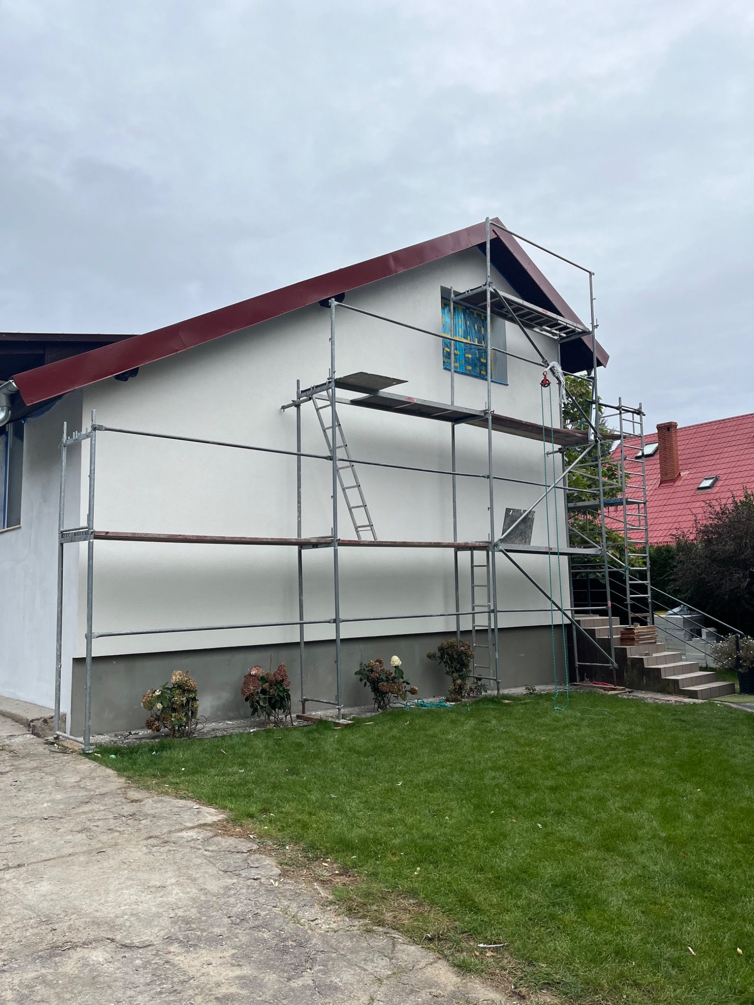 Dom jednorodzinny w trakcie prac elewacyjnych; widoczne rusztowanie, częściowo pomalowana ściana na biało, okno zabezpieczone folią, czerwony dach.