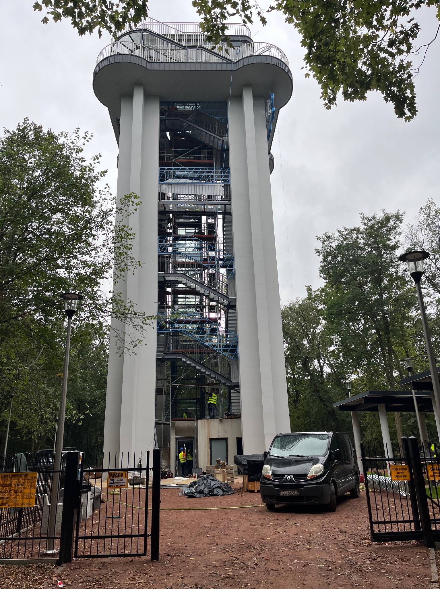 Wysoka wieża widokowa w trakcie prac wykończeniowych, widoczne rusztowania i pracownicy w odblaskowych kamizelkach, na pierwszym planie czarny van i ogrodzenie z tablicami informacyjnymi.