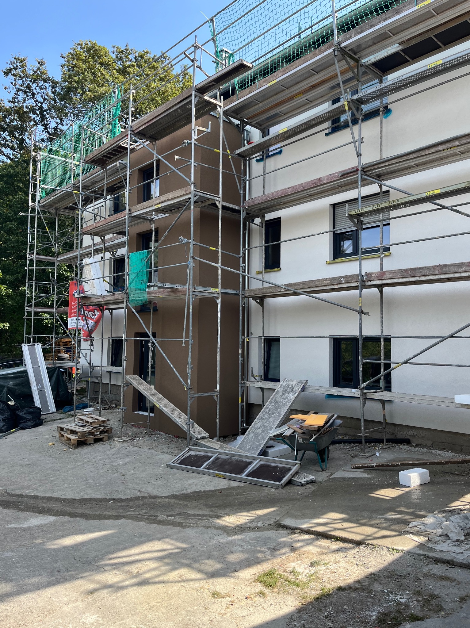 Budynek wielorodzinny w trakcie prac elewacyjnych; widoczne rusztowania, fragment ocieplonej i otynkowanej ściany w kolorze brązowym oraz biała ściana po tynkowaniu, z czarnymi oknami; na ziemi...