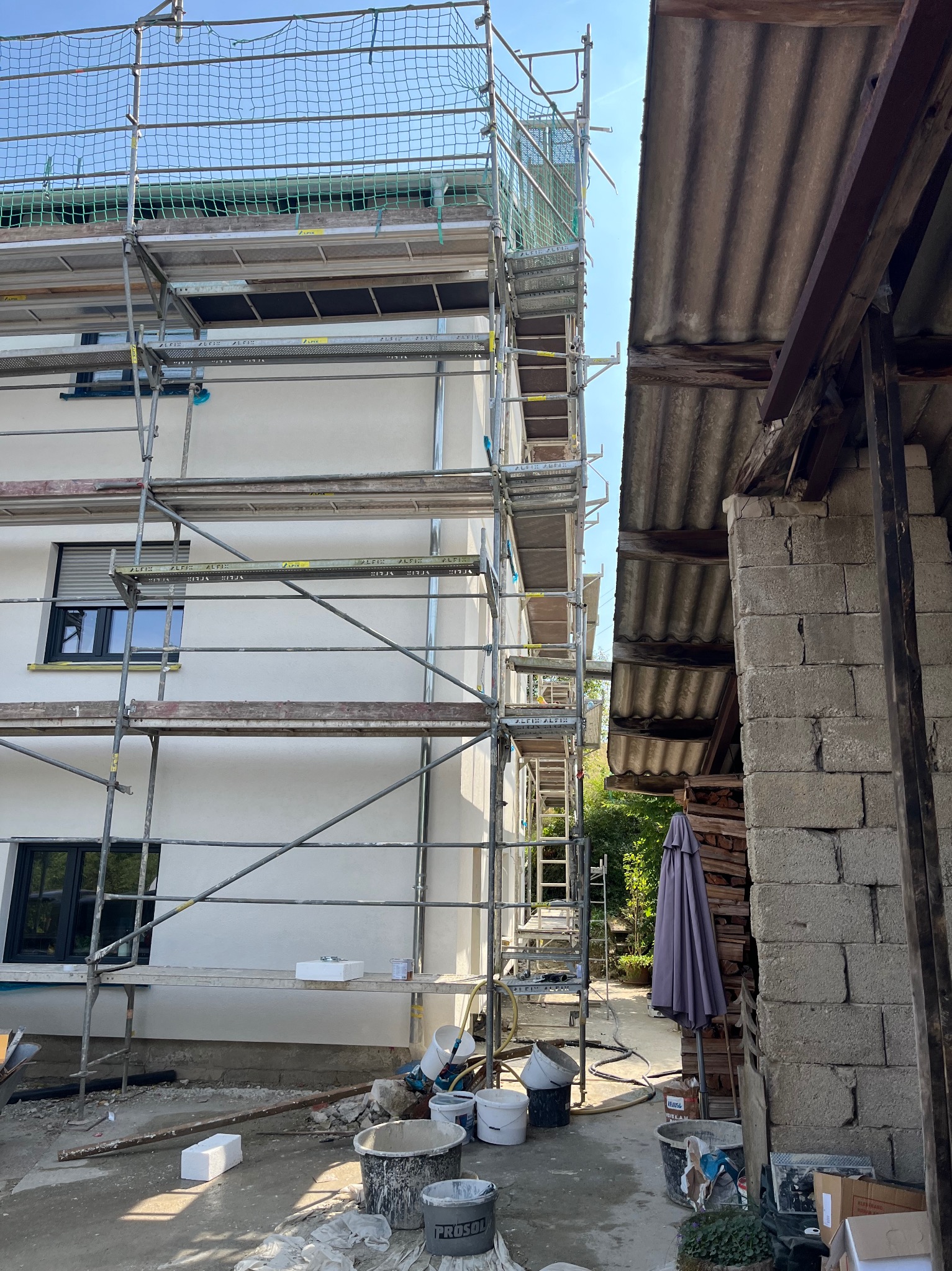 Elewacja budynku w trakcie prac wykończeniowych, widoczne rusztowania, wiadra z zaprawą i materiały izolacyjne.