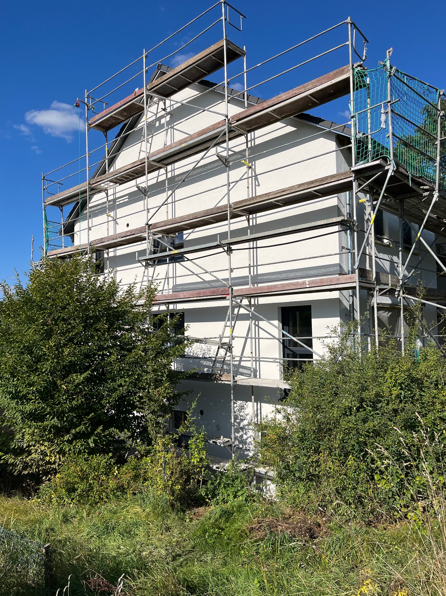 Dom jednorodzinny w trakcie prac elewacyjnych, otoczony rusztowaniem i bujną zielenią, widoczne świeżo otynkowane ściany w jasnym odcieniu.