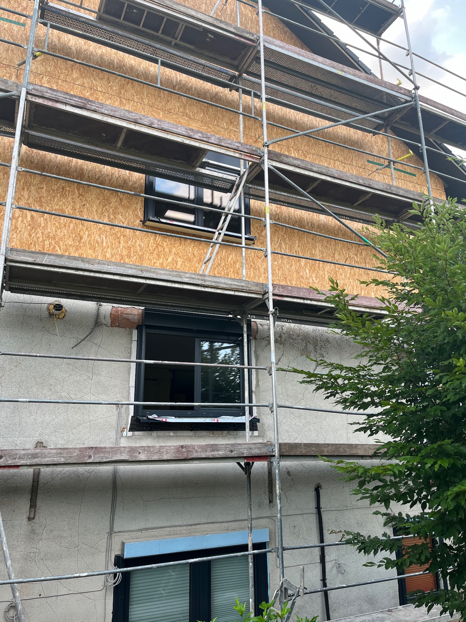 Dom w trakcie ocieplania elewacji, widoczne rusztowanie, płyty OSB na ścianach i okna z czarnymi ramami.