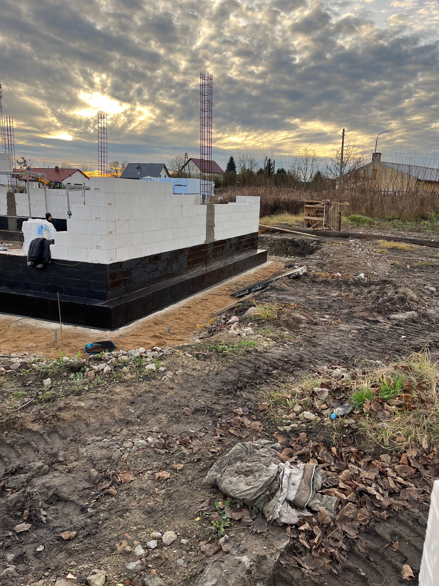 Budowa domu jednorodzinnego w trakcie wznoszenia ścian z bloczków betonowych, izolacja fundamentów czarną masą bitumiczną, stalowe pręty zbrojeniowe wystające z fundamentów, tło: pochmurne niebo...