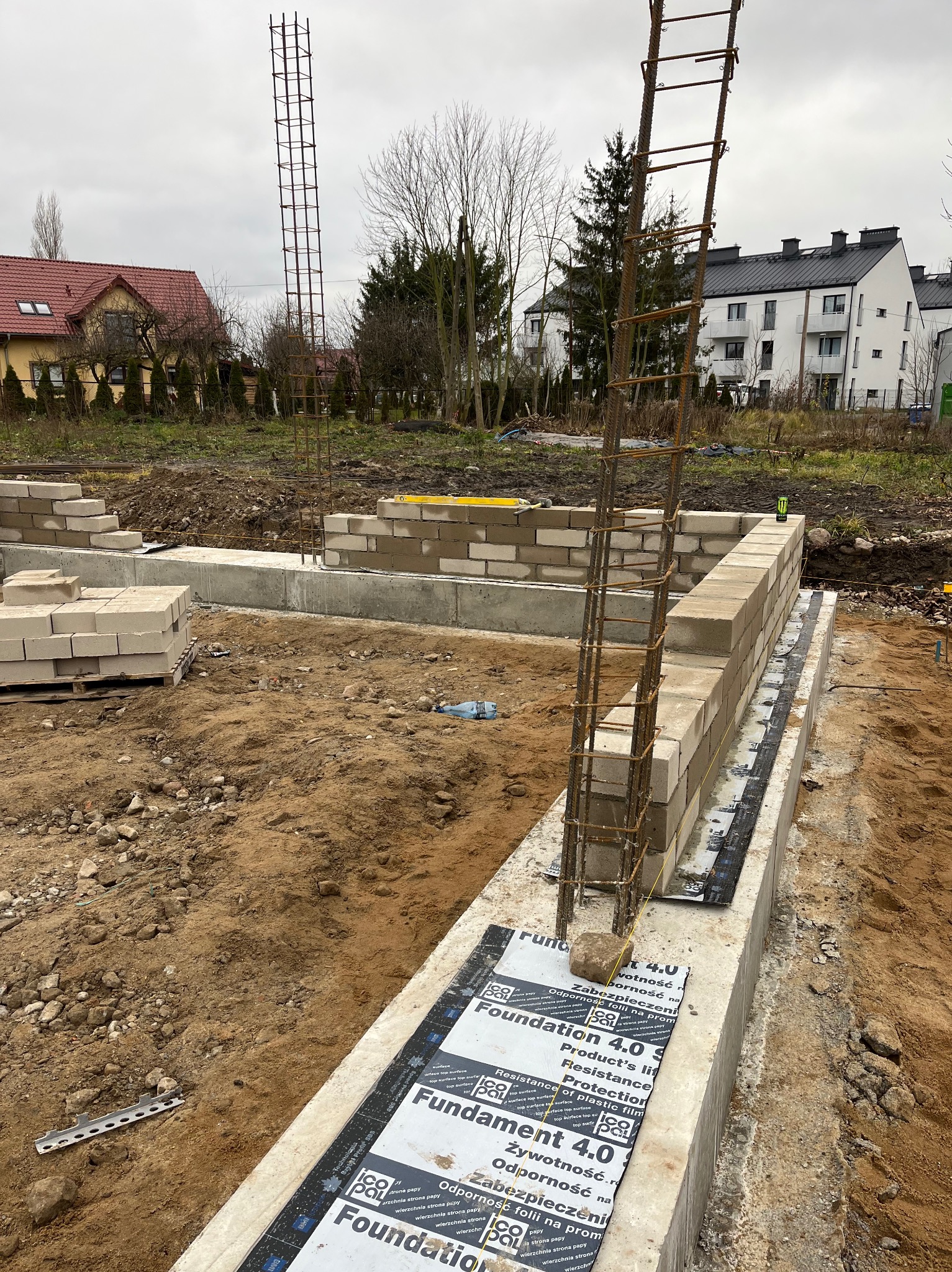 Budowa fundamentów domu jednorodzinnego: betonowy fundament z hydroizolacją, bloczki betonowe, zbrojenie pionowe, teren budowy z widocznymi domami w tle.