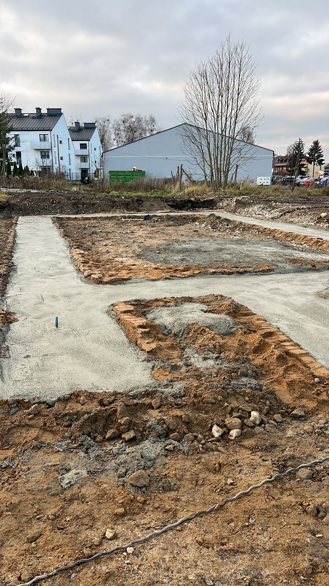 Przygotowanie fundamentów pod budynek, widoczne wykopane i zalane betonem ławy fundamentowe, zarys przyszłego budynku, w tle domy jednorodzinne i pochmurne niebo.