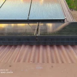 Panele fotowoltaiczne zamontowane na pofałdowanym, pomarańczowo-brązowym dachu z widocznymi śladami zabrudzeń i zacieków, uchwycone z niskiego kąta.