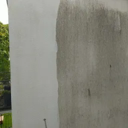 Częściowo umyta elewacja budynku z widoczną różnicą czystości, strumień wody z lancy myjącej w dolnej części kadru. Widok z dołu, fragment balkonu w tle.