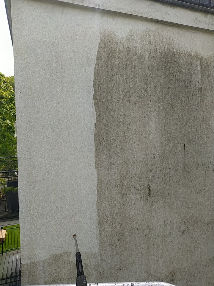 Częściowo umyta elewacja budynku z widoczną różnicą czystości, strumień wody z lancy myjącej w dolnej części kadru. Widok z dołu, fragment balkonu w tle.