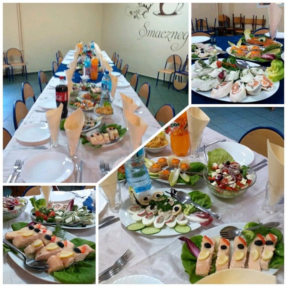 Długi stół nakryty białym obrusem, z różnorodnymi przekąskami, napojami i zastawą, w tle napis 'Smacznego' na ścianie i krzesła