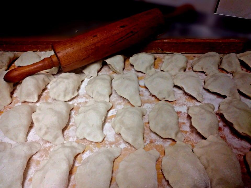 Ręcznie lepione pierogi ułożone na drewnianej desce posypanej mąką, obok leży drewniany wałek.