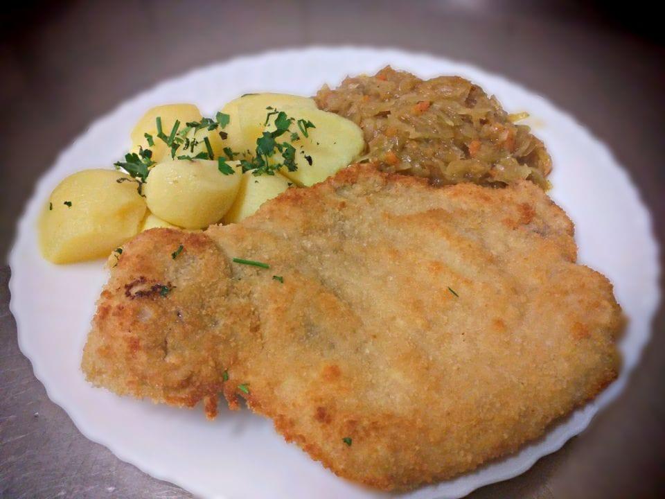 Kotlet schabowy w panierce z ziemniakami posypanymi koperkiem i kapustą duszoną na białym talerzu.