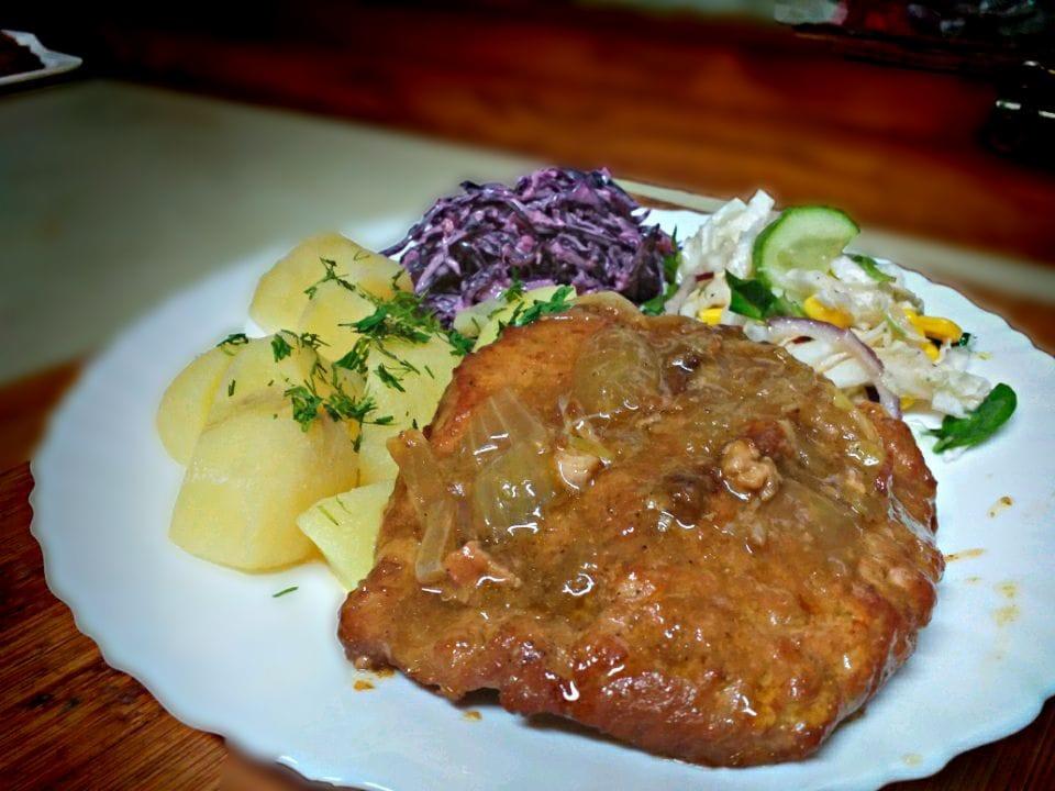Kotlet w sosie z cebulą podany z ziemniakami posypanymi koperkiem, surówką z czerwonej kapusty i sałatką z kukurydzy, ogórka i cebuli na białym talerzu z karbowanym brzegiem.