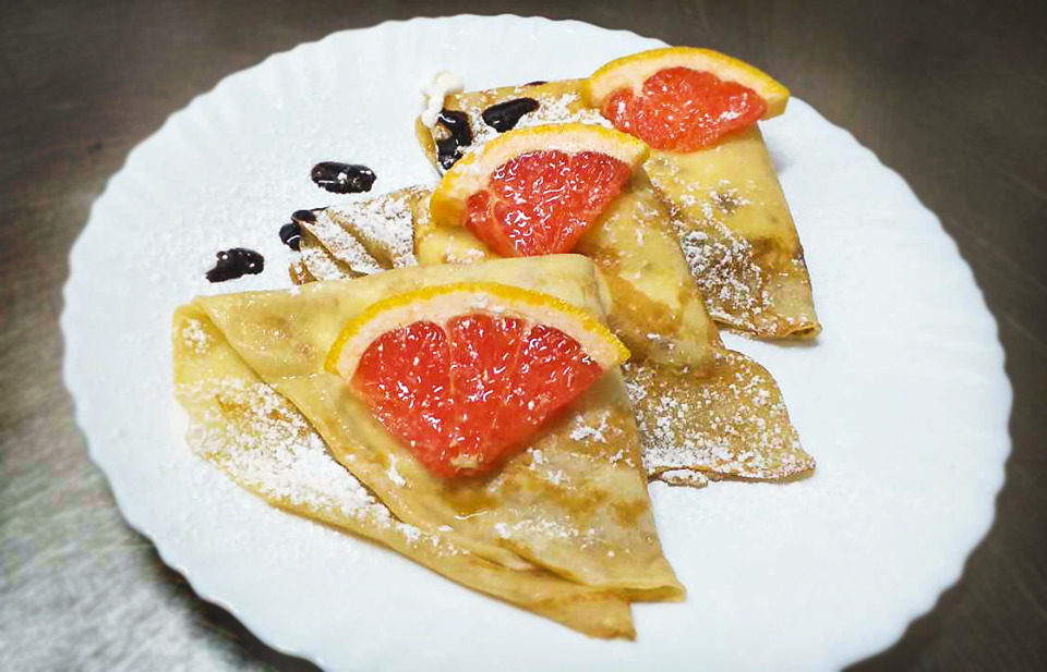 Delikatne naleśniki polane sosem czekoladowym i posypane cukrem pudrem, udekorowane soczystymi cząstkami grejpfruta na białym talerzu.