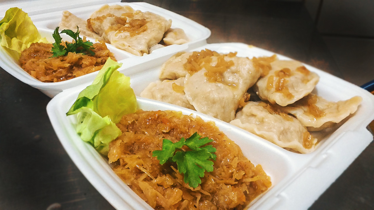 Dwa białe, styropianowe pojemniki z pierogami polanymi podsmażoną cebulką i kapustą z grzybami, udekorowane zieloną sałatą i natką pietruszki.