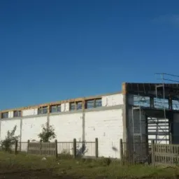 Budowa hali przemysłowej z białych bloczków, widoczne rusztowania i konstrukcja dachu, niebieskie niebo w tle.