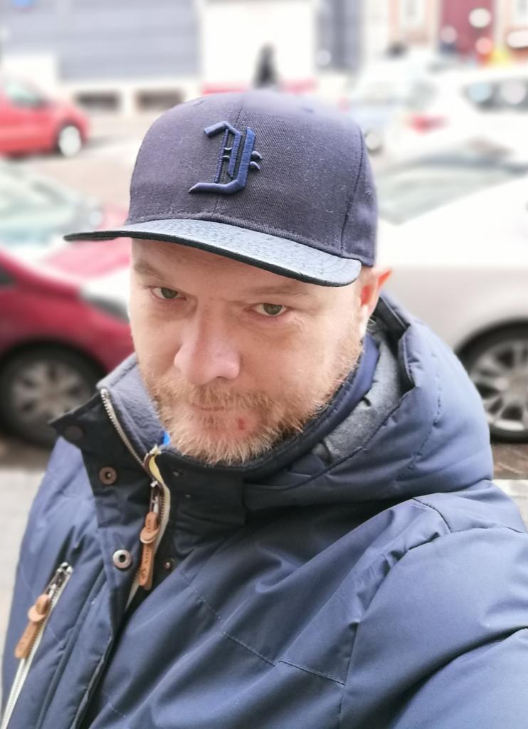 Portret mężczyzny w niebieskiej kurtce i czapce z literą 'D' na tle parkingu z samochodami.