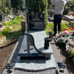 Nowo postawiony, jasnoszary pomnik z czarną tablicą nagrobną, krzyżem i portretem zmarłej osoby, otoczony świeżymi kwiatami i zniczami, na tle cmentarza z drzewami i innymi nagrobkami, z mężczyzną...