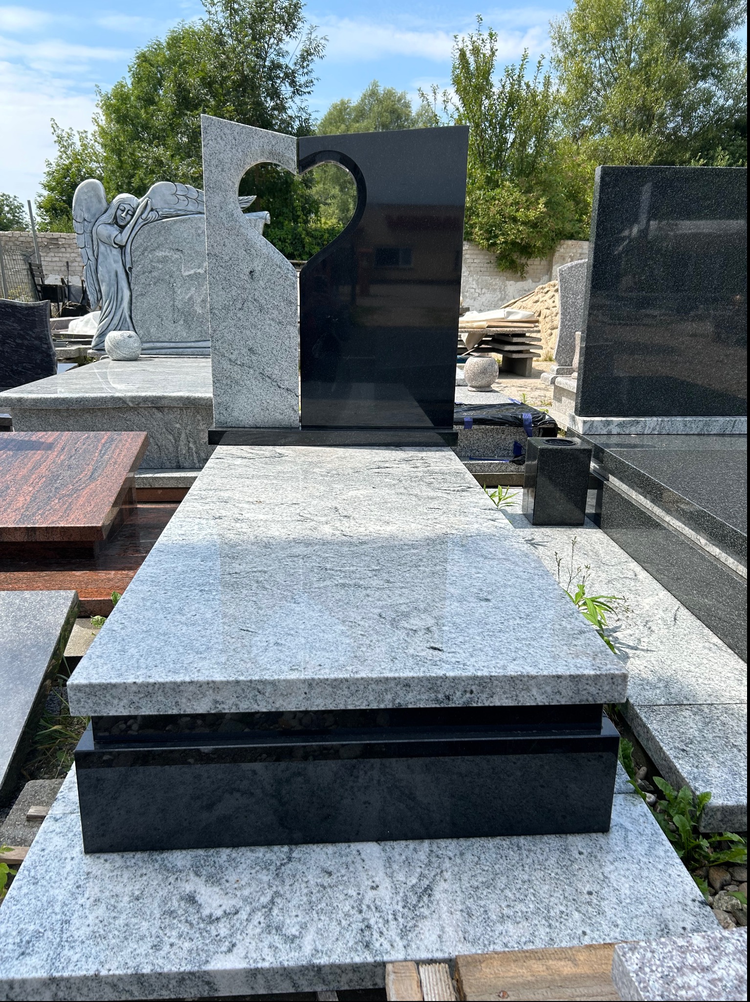 Nowoczesny, jasnoszary pomnik z czarnym elementem w kształcie serca, otoczony zielenią i innymi nagrobkami.