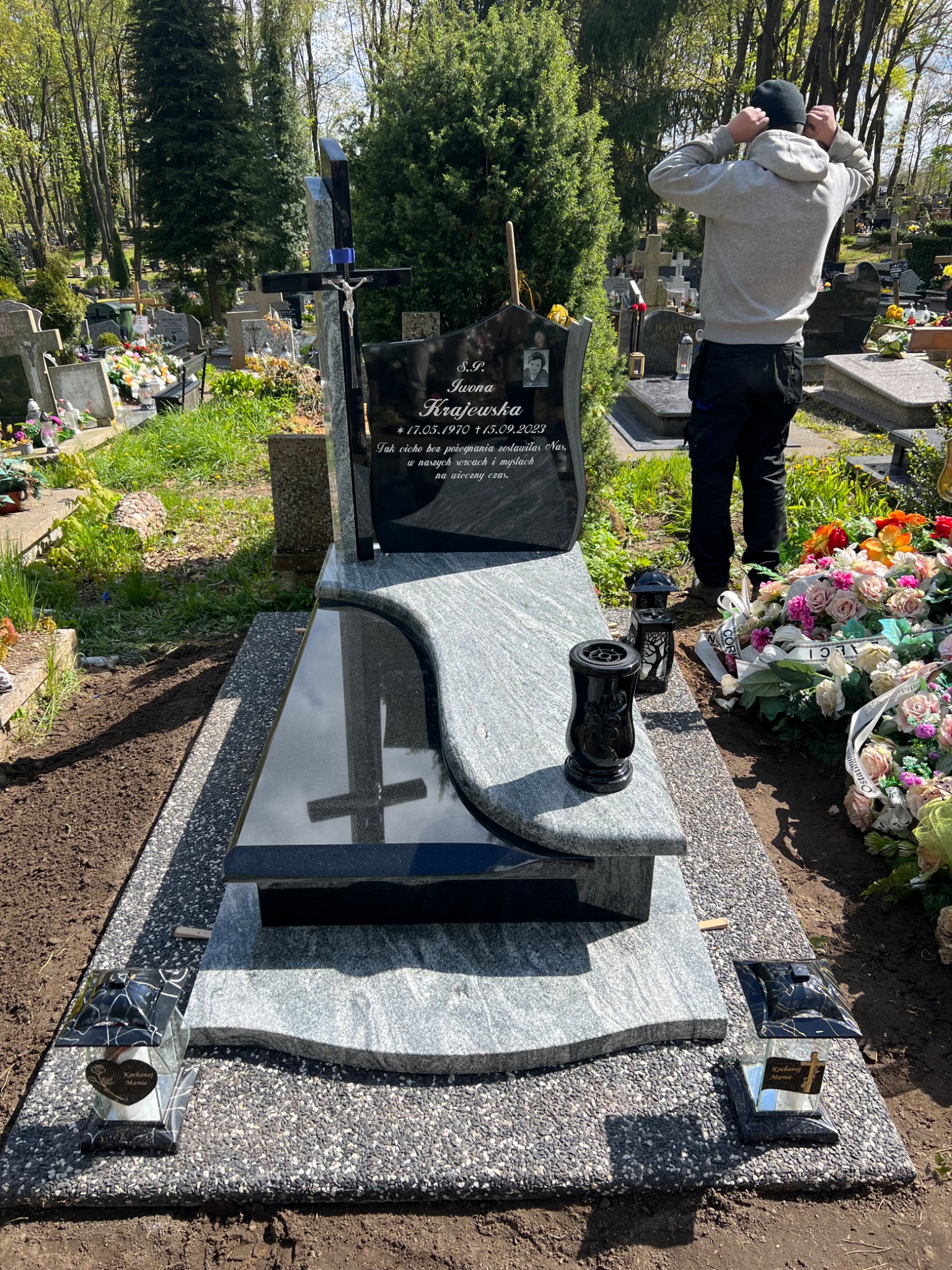 Nowo postawiony, jasnoszary pomnik z czarną tablicą nagrobną, krzyżem i portretem zmarłej osoby, otoczony świeżymi kwiatami i zniczami, na tle cmentarza z drzewami i innymi nagrobkami, z mężczyzną...