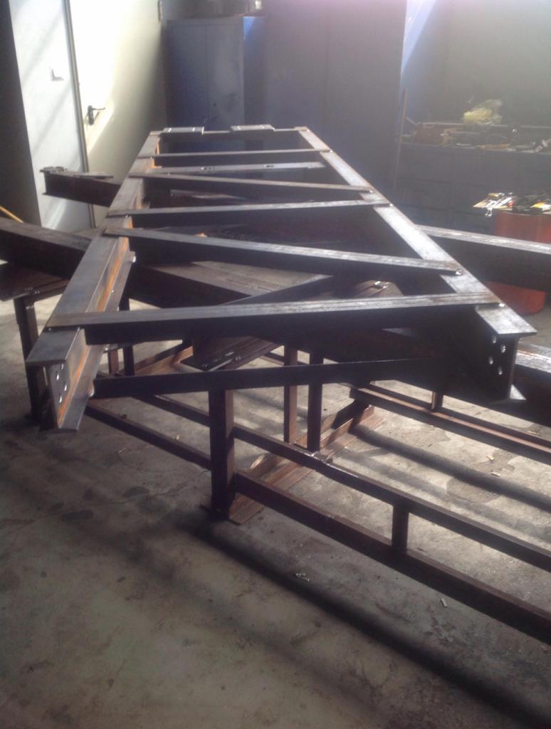 Stalowa konstrukcja szkieletowa z profili spawanych, widoczne spawy, surowy metal, szara podłoga warsztatowa, oświetlenie dzienne.