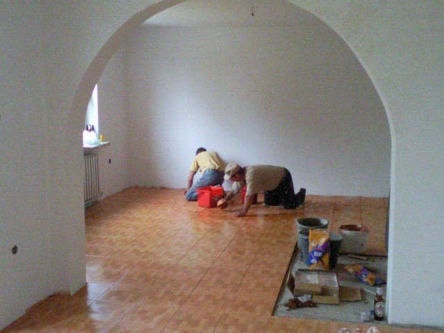 Dwóch pracowników układa brązowe płytki podłogowe w pomieszczeniu z białymi ścianami i łukowym przejściem, widoczne narzędzia i materiały budowlane.