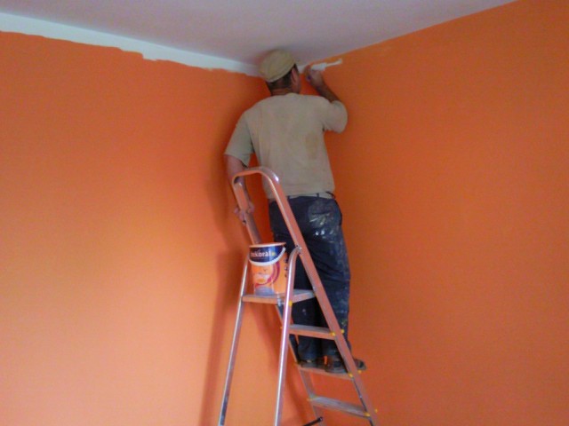 Mężczyzna na aluminiowej drabinie maluje pomarańczową ścianę na biało przy suficie, widoczna puszka farby marki Dekoral.