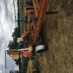 Pomarańczowa, przenośna traka taśmowa do drewna na łące z pociętymi kłodami w pobliżu, pochmurne niebo w tle.