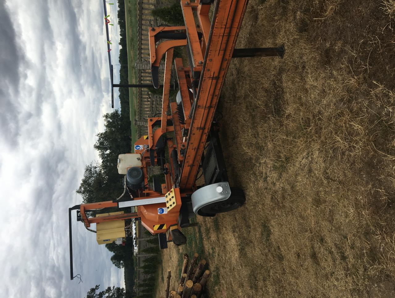 Pomarańczowa, przenośna traka taśmowa do drewna na łące z pociętymi kłodami w pobliżu, pochmurne niebo w tle.