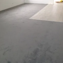 Szara wykładzina dywanowa w pomieszczeniu, częściowo ułożona, z widocznym fragmentem podłogi z paneli w tle, sugerującym etap prac wykończeniowych.