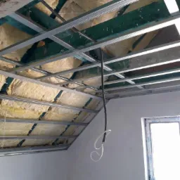 Skośny sufit poddasza w trakcie ocieplania wełną mineralną, widoczne metalowe profile konstrukcyjne i przewód elektryczny. Okno w tle.