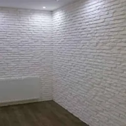 Ściana wewnętrzna wykończona białą, dekoracyjną cegłą, częściowo ułożoną, obok grzejnik i drewnopodobna podłoga.