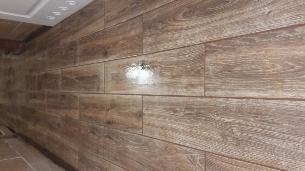 Ułożone, brązowe panele imitujące drewno na ścianie, z widoczną fugą, ułożone poziomo z perspektywicznym skrótem. Widoczne oświetlenie sufitowe.