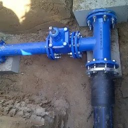 Niebieski, żeliwny trójnik z kołnierzami i zaworem, część podziemnego przyłącza wodociągowego, wsparty na betonowych blokach w wykopie ziemnym.