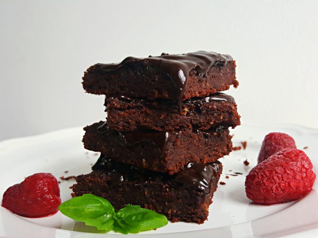 Stos brązowych, błyszczących brownie polanych czekoladą, udekorowany malinami i listkiem bazylii na białym talerzu.