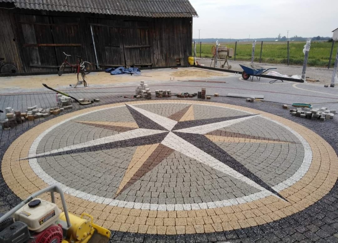 Kompas z kostki brukowej ułożony na podwórku, widoczna ubijarka, sterta kostki i stara stodoła w tle.