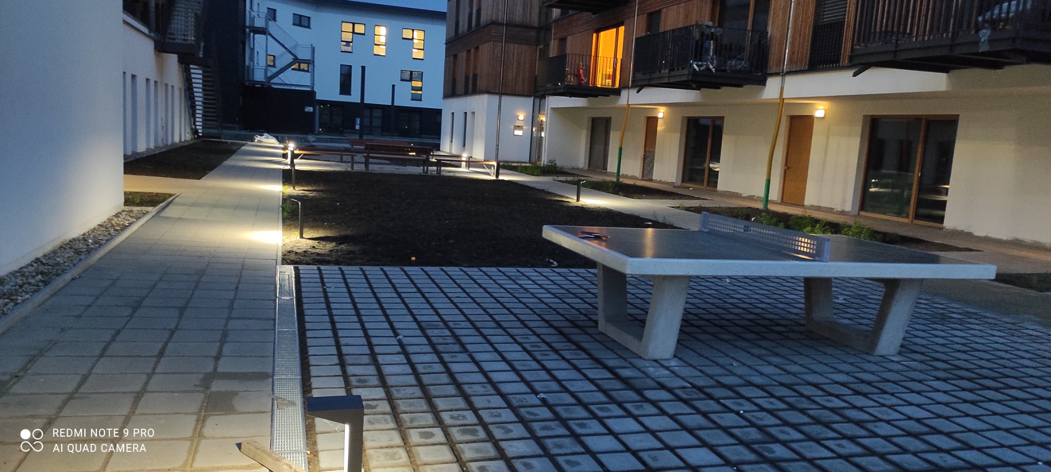 Nowoczesna przestrzeń osiedlowa z betonową kostką, oświetlonymi ścieżkami i stołem do ping-ponga, na tle budynków mieszkalnych oświetlonych wieczorem.