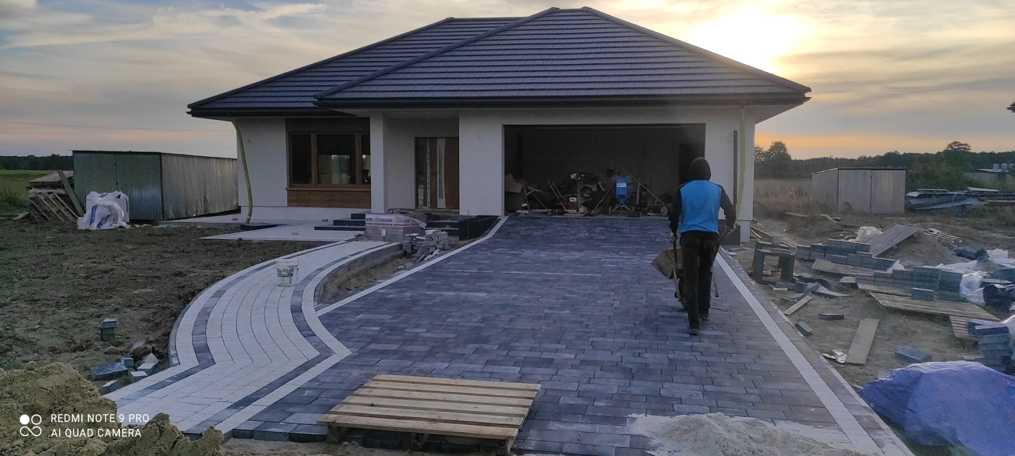 Nowoczesny podjazd z kostki brukowej w trakcie układania, widoczny pracownik z taczką, dom jednorodzinny w tle, paleta z kostką i narzędzia budowlane.