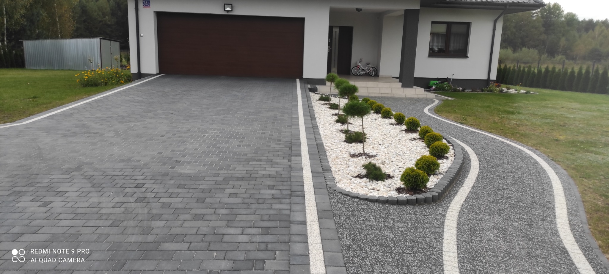 Nowoczesny podjazd do domu z szarej kostki brukowej, wykończony białym kamieniem i ozdobnymi roślinami, z widoczną bramą garażową w tle.