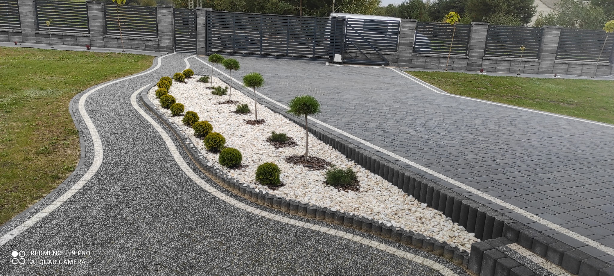 Ścieżka z kostki brukowej w odcieniach szarości, oddzielona białym pasem od trawnika, z ozdobnym rabatem wypełnionym białym kamieniem i niskimi krzewami kulistymi oraz miniaturowymi drzewkami...