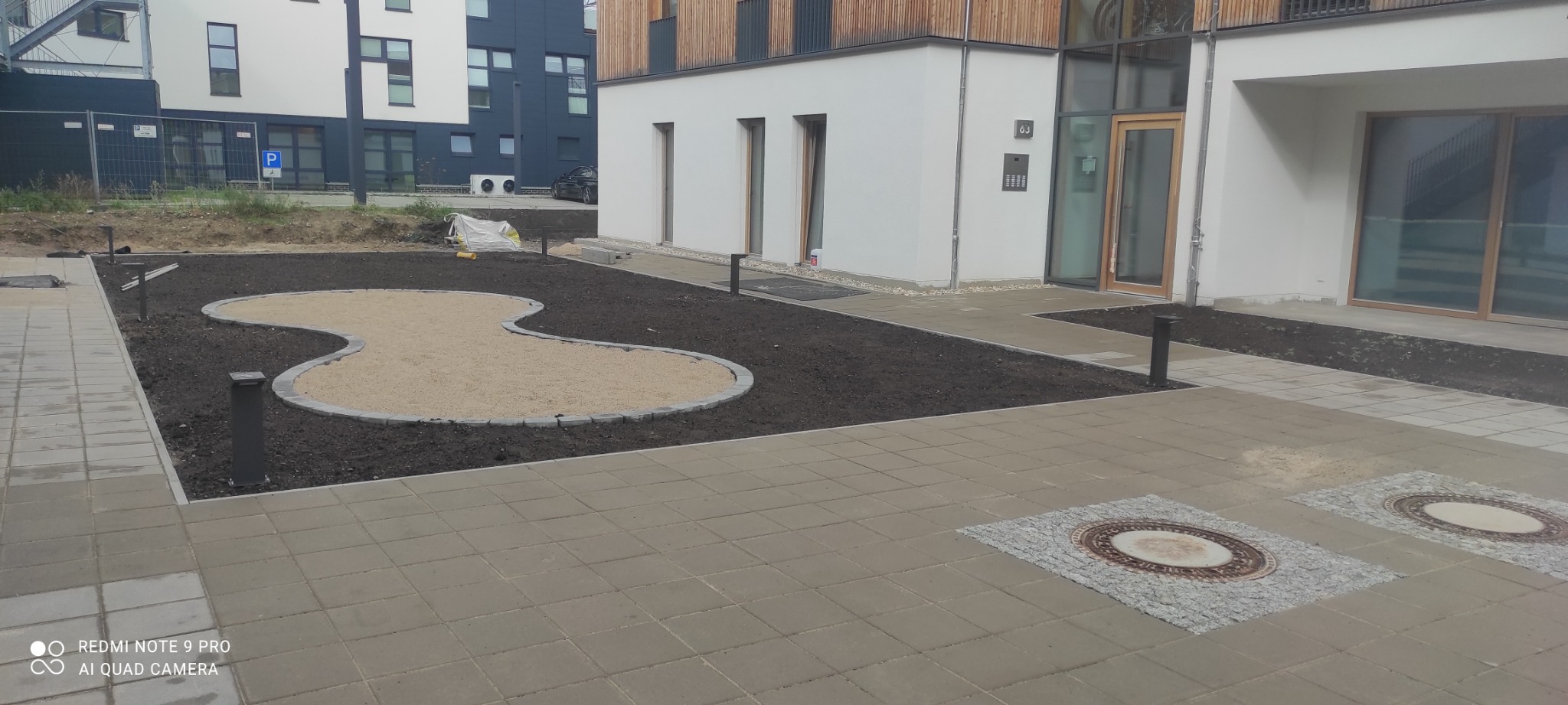 Ułożona kostka brukowa przed nowoczesnym budynkiem, z wydzielonym obszarem ziemi przygotowanym pod nasadzenia i piaskownicą o nieregularnym kształcie, oświetloną niskimi lampami.