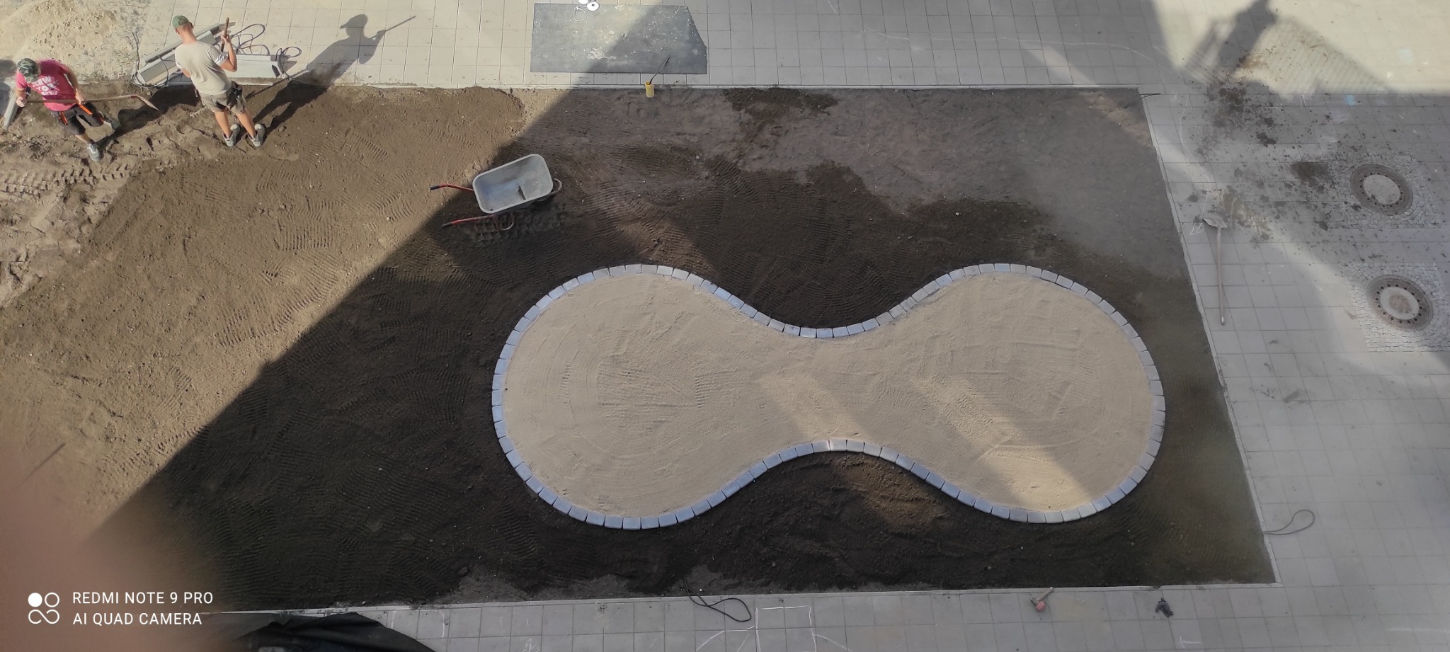 Z góry widok na teren przygotowywany pod nawierzchnię z piasku, obrzeża z kostki ułożone w kształt ósemki, dwóch pracowników rozrzuca ziemię, obok stoi taczka.
