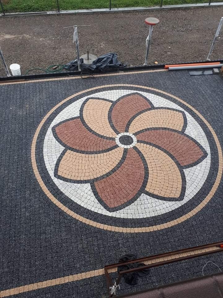 Ozdobna nawierzchnia z kostki brukowej ułożona w motyw kwiatowy z ciemnoszarym tłem i elementami w kolorach beżowym, brązowym i białym, widok z góry.