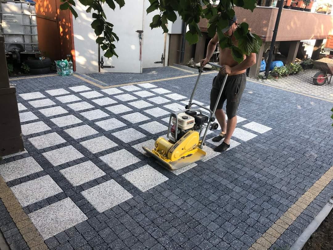 Pracownik zagęszcza wibroprasa wzór z kostki brukowej w szachownicę z obramowaniem z kostki w kolorze piaskowym na podjeździe przed budynkiem.