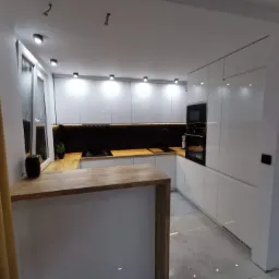 Nowoczesna, minimalistyczna kuchnia z białymi, lakierowanymi szafkami, drewnianym blatem i czarną mozaiką nad blatem, oświetlona punktowymi lampami sufitowymi, widok zza zasłony