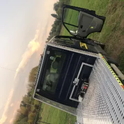 Błękitna laweta z otwartymi drzwiami kabiny, zaparkowana na trawiastym terenie z widocznymi drzewami w tle i zachodzącym słońcem.