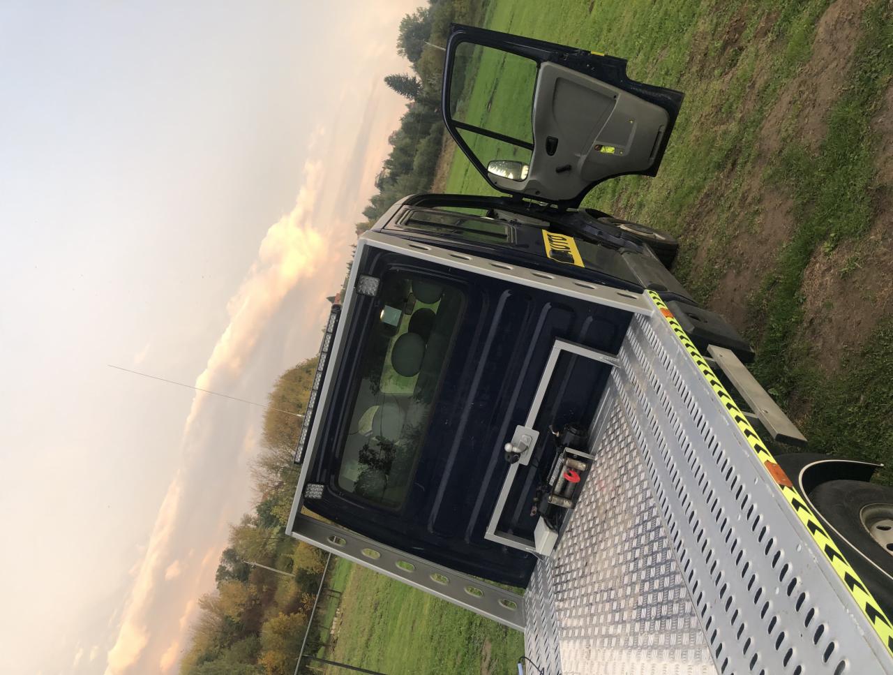 Błękitna laweta z otwartymi drzwiami kabiny, zaparkowana na trawiastym terenie z widocznymi drzewami w tle i zachodzącym słońcem.